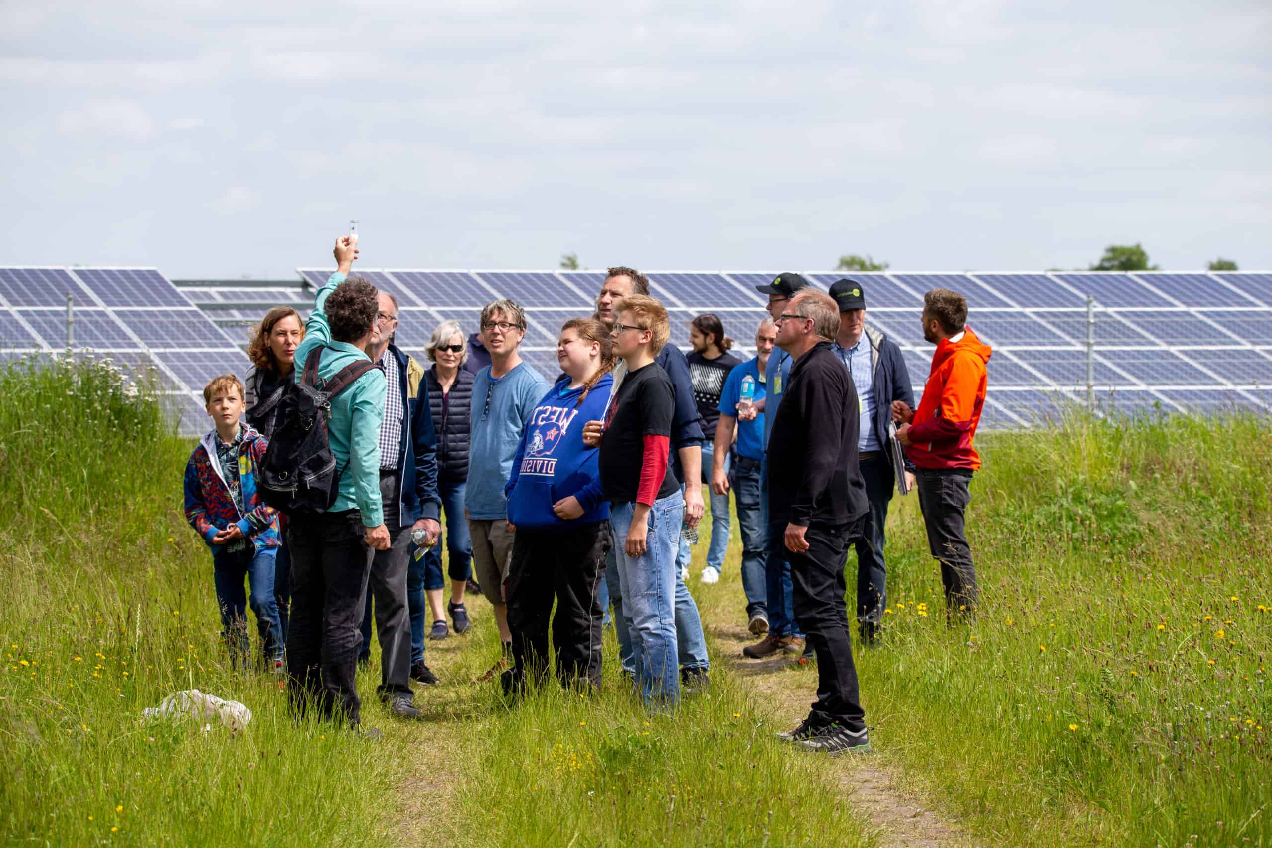 Gruppe von Menschen unterschiedlichen Alters in einem Solarpark. Eine Person hält ein Objektivglas in die Luft und erklärt der Gruppe etwas. Im Hintergrund sieht man den Solarpark.